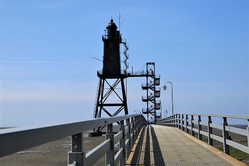 A wooden bridge leading to a metal lighthouse overlooking the water.