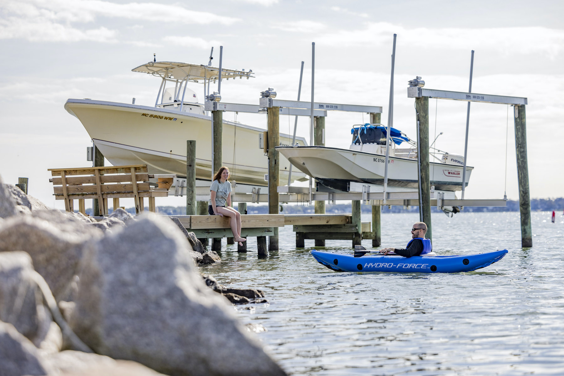 Man kayaking with woman sitting on pier with boats near the pier.