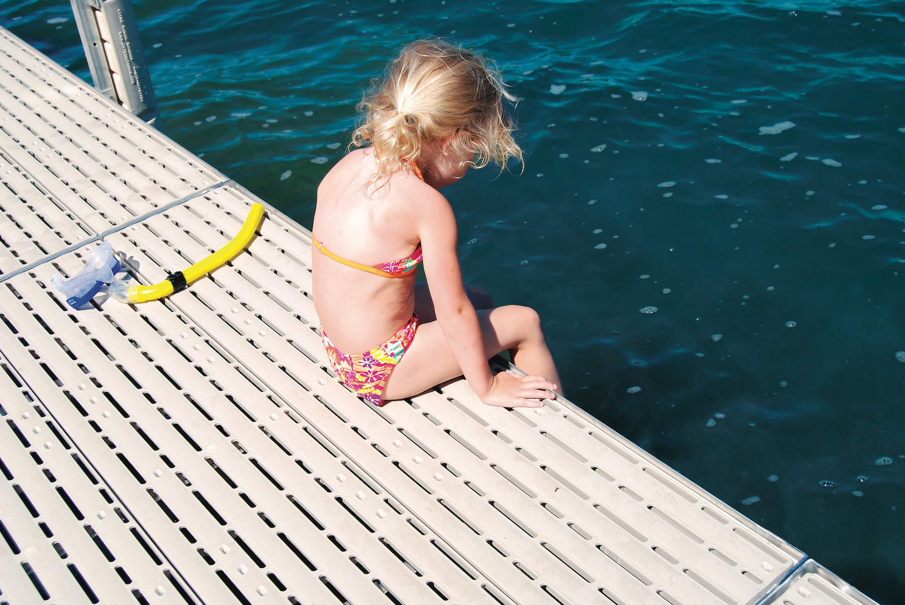 A young girl sitting at the edge of a dock looking into the blue water.