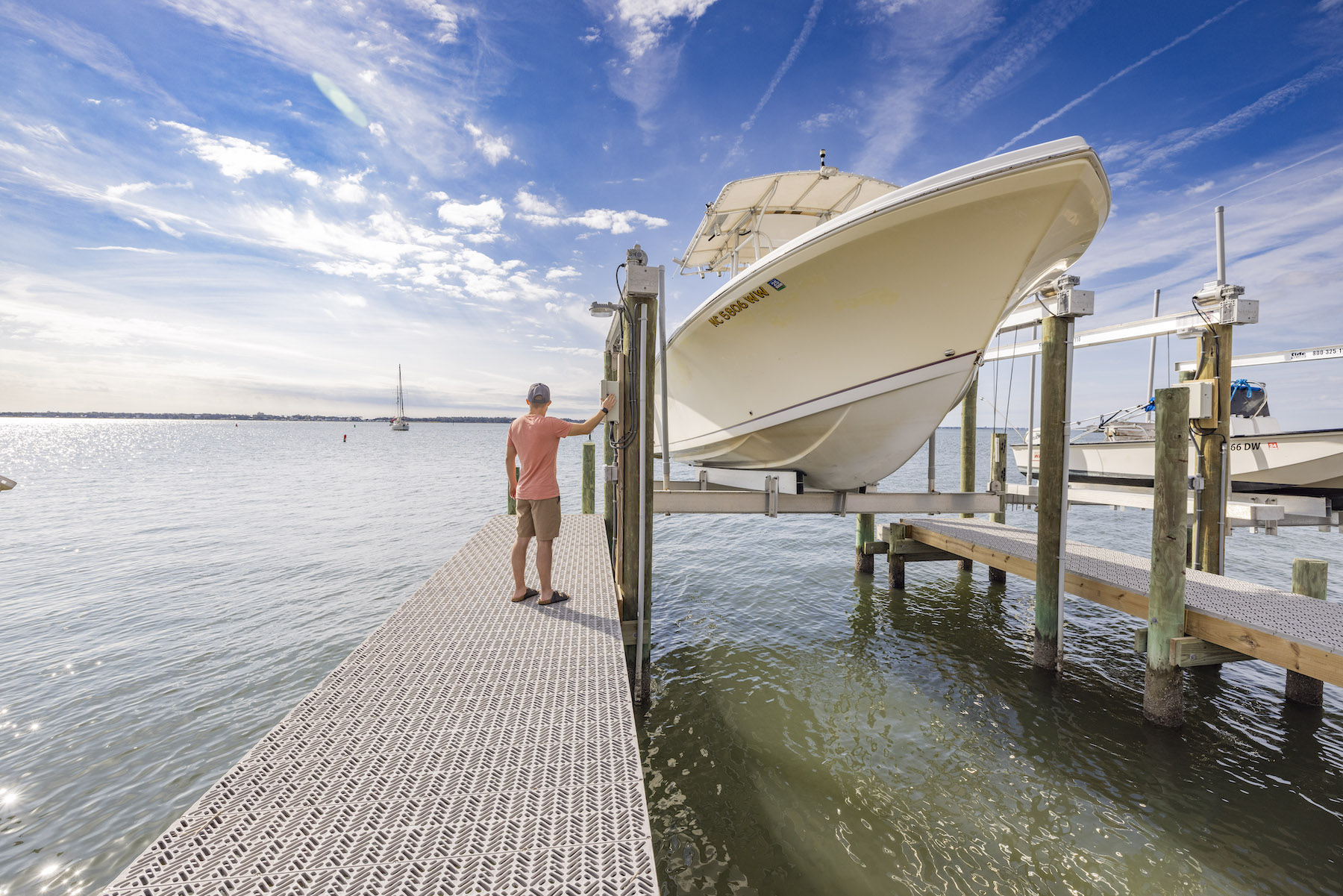 A person standing on a dock, next to a boat raised in a boat lift. The person is inspecting the boat lift’s electrical box.