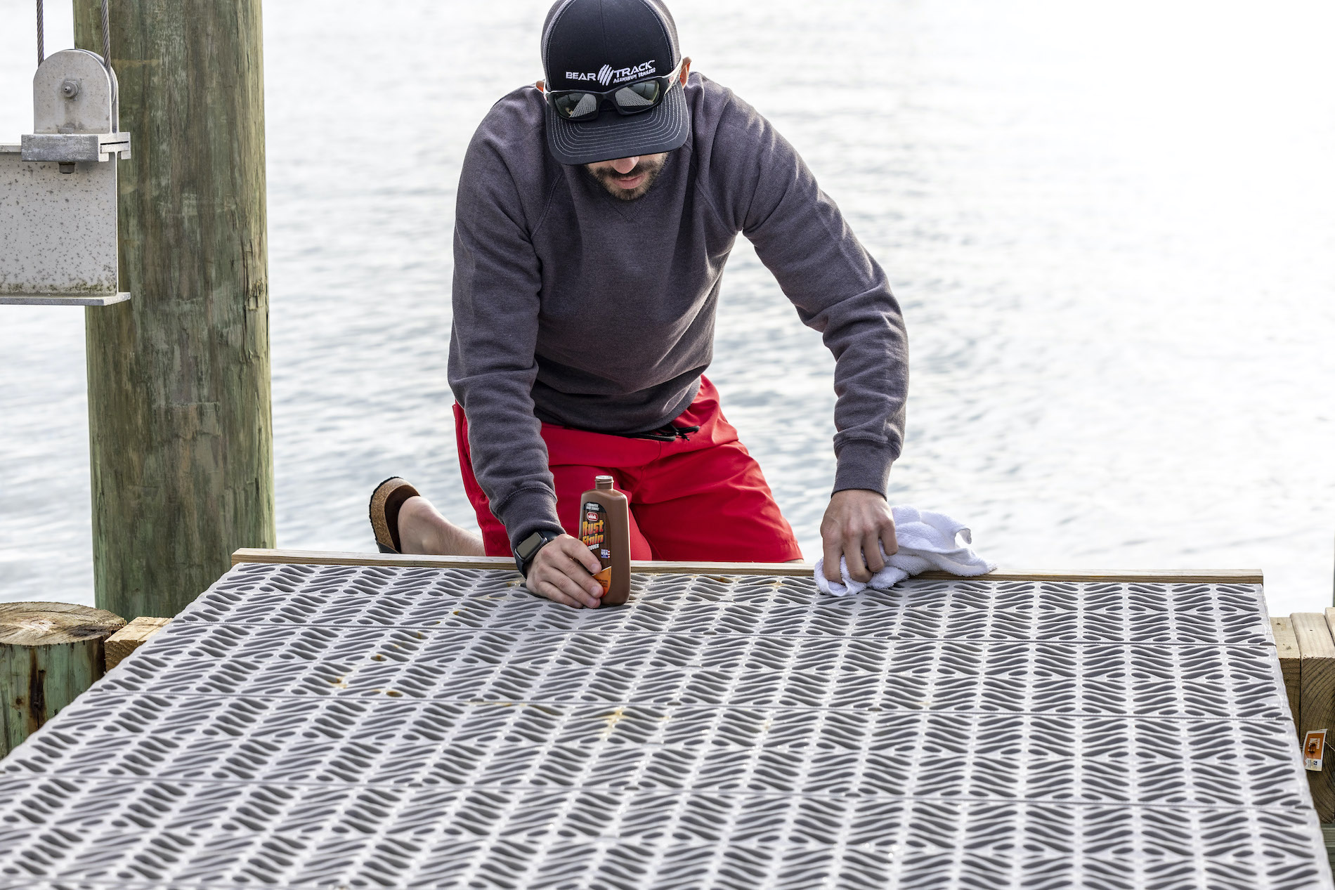 A person is kneeling next to a dock and is applying cleaning product with a rag to the dock. The person is wearing a hat with sunglasses on top, red shorts, and a gray long-sleeve shirt.