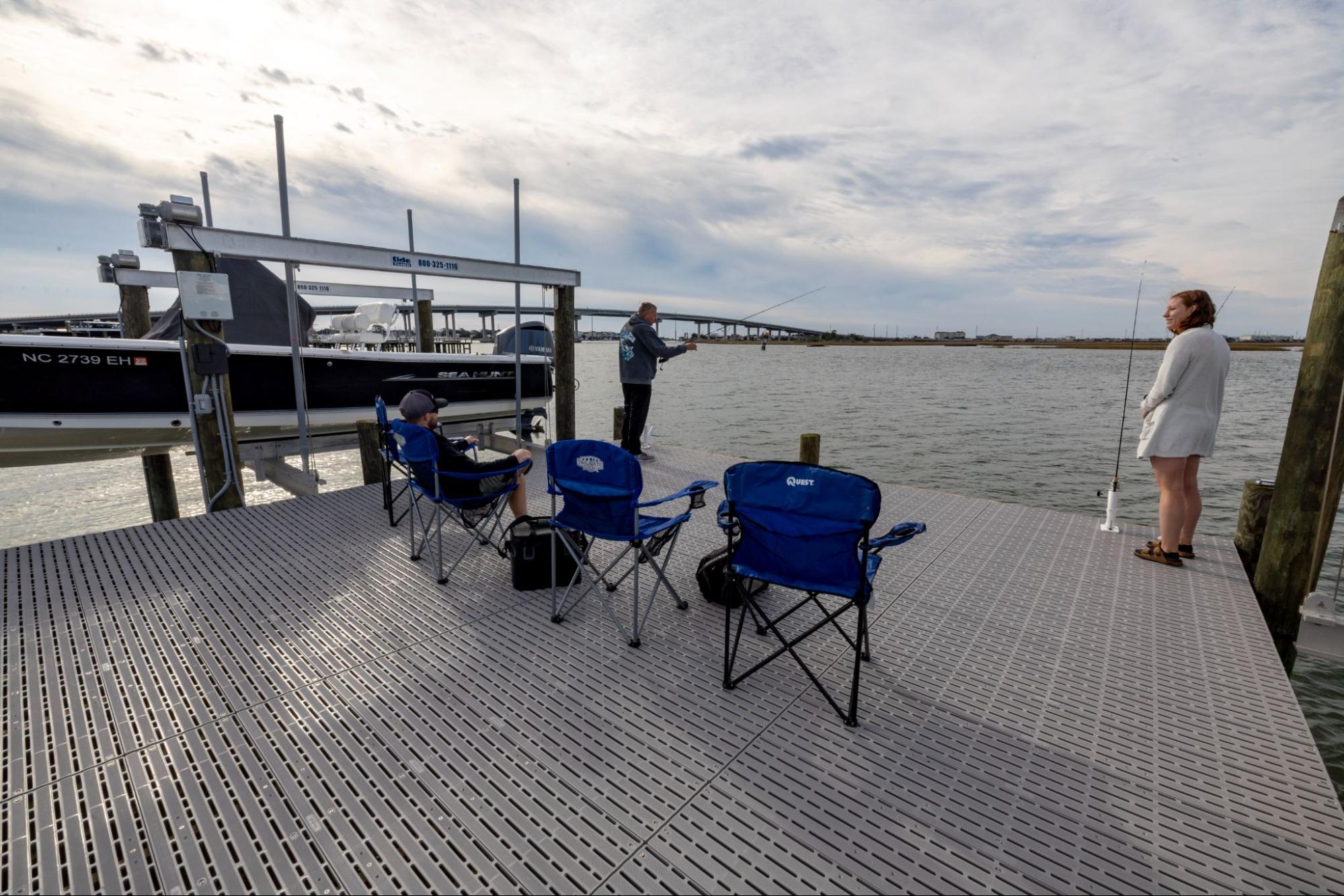 Three people stand on a marine dock with Titan Deck decking. One is holding a fishing pole and one is sitting in one of three blue folding chairs.