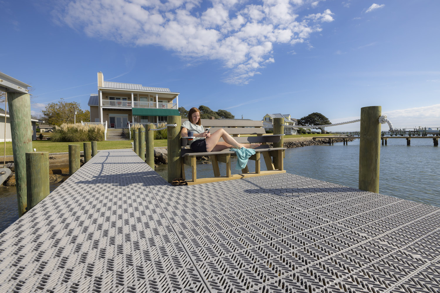 There is a person sitting on a dock bench looking at their phone. There is a house behind the person at the base of the dock and water surrounds the dock.