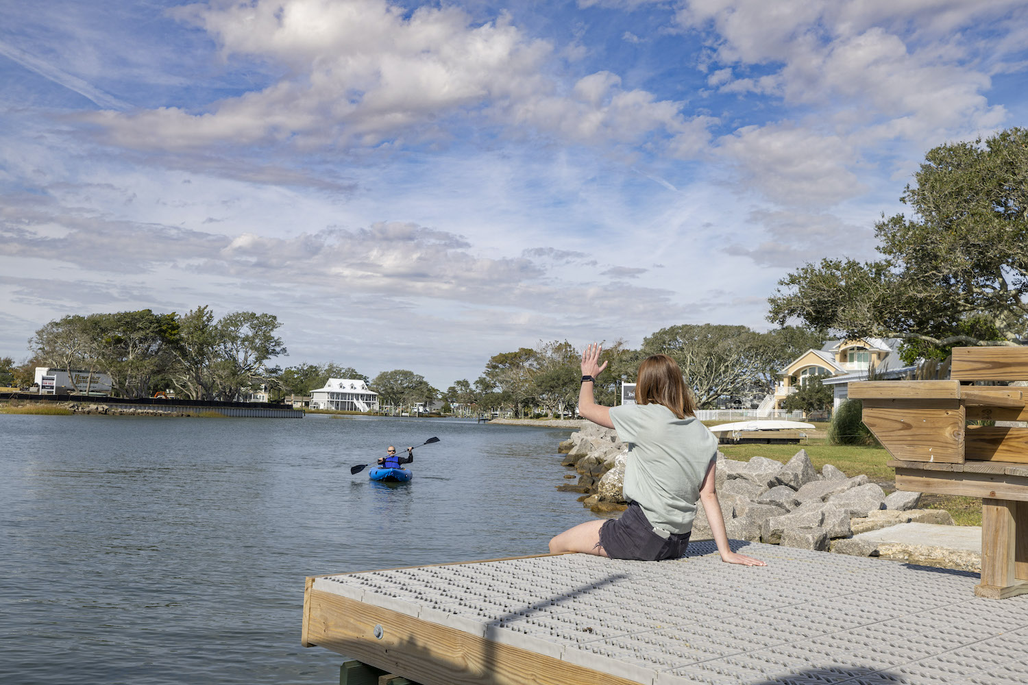 A person in a light green shirt and black shorts is waving to another person paddling a kayak on the lake. 