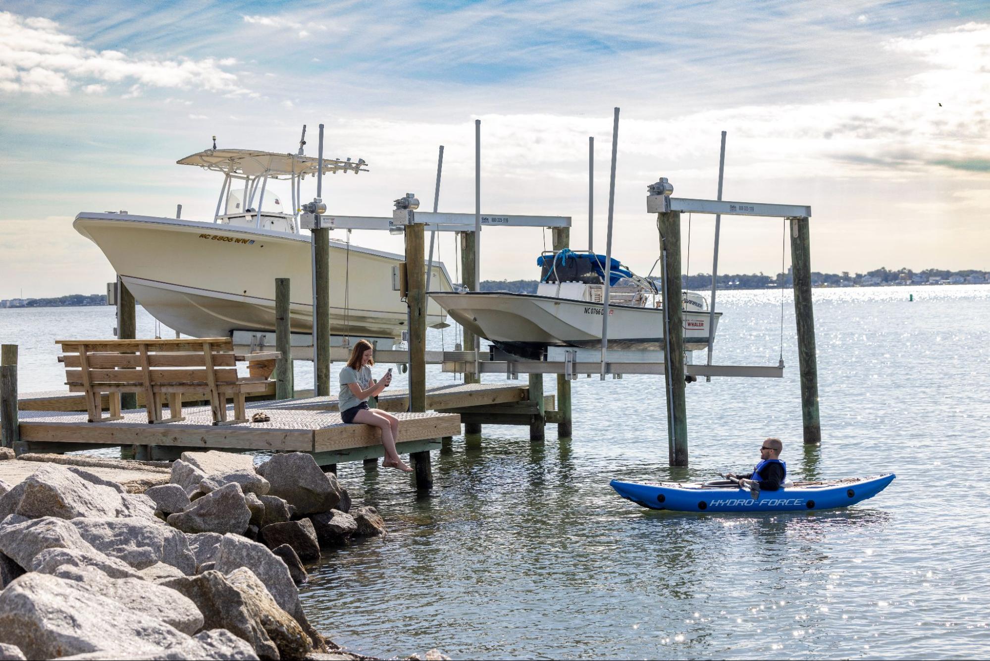 An image of one person sitting on a dock while another person paddles towards them in a kayak. 