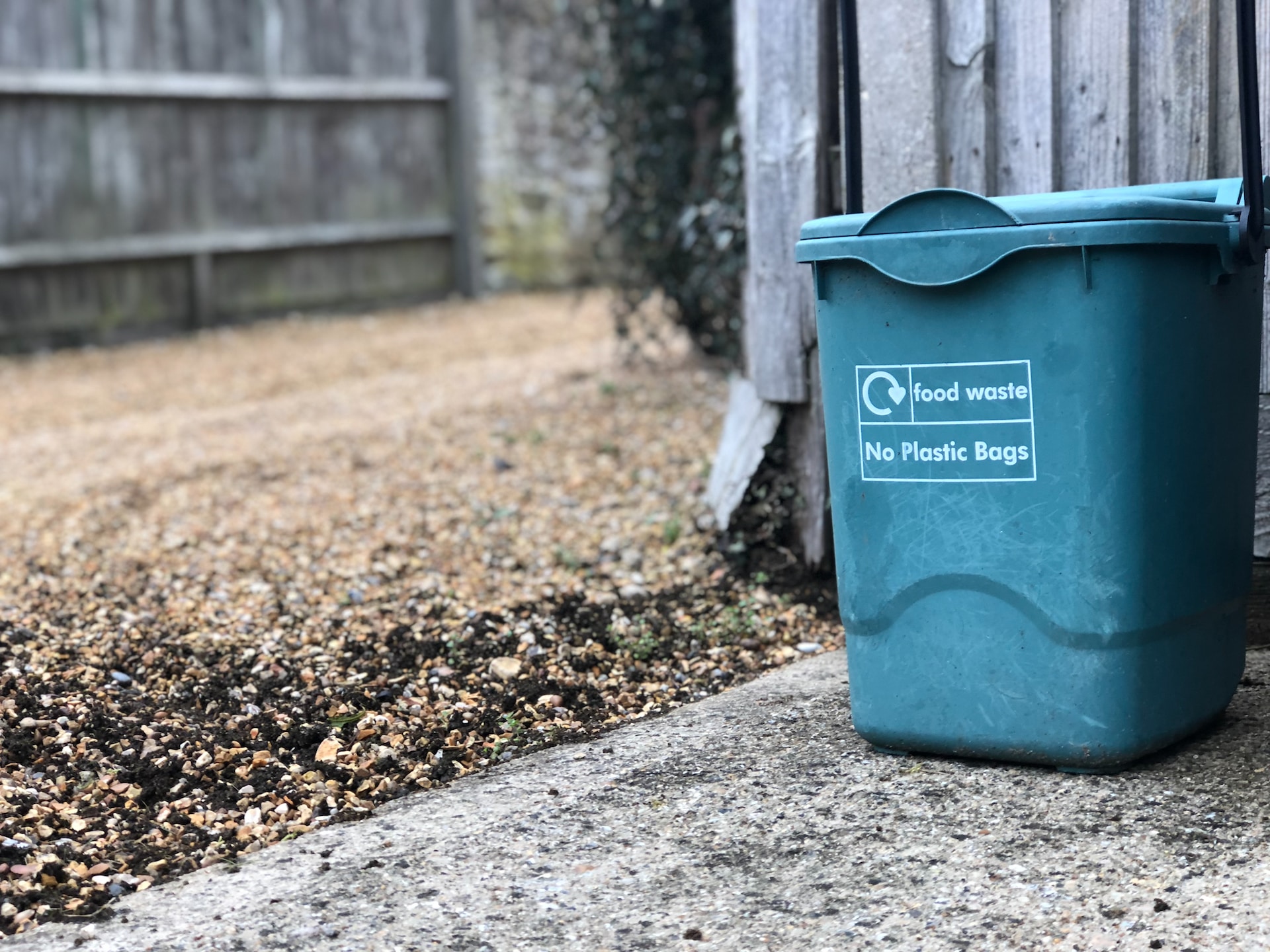 An image of an outdoor, dark green food waste bin. The bin is set on concrete and next to a yard.