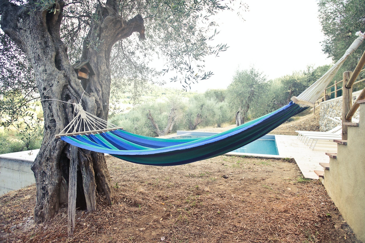 A blue striped hammock hanging between a tree and an outdoor stair railing. There is an outdoor pool behind the hammock.