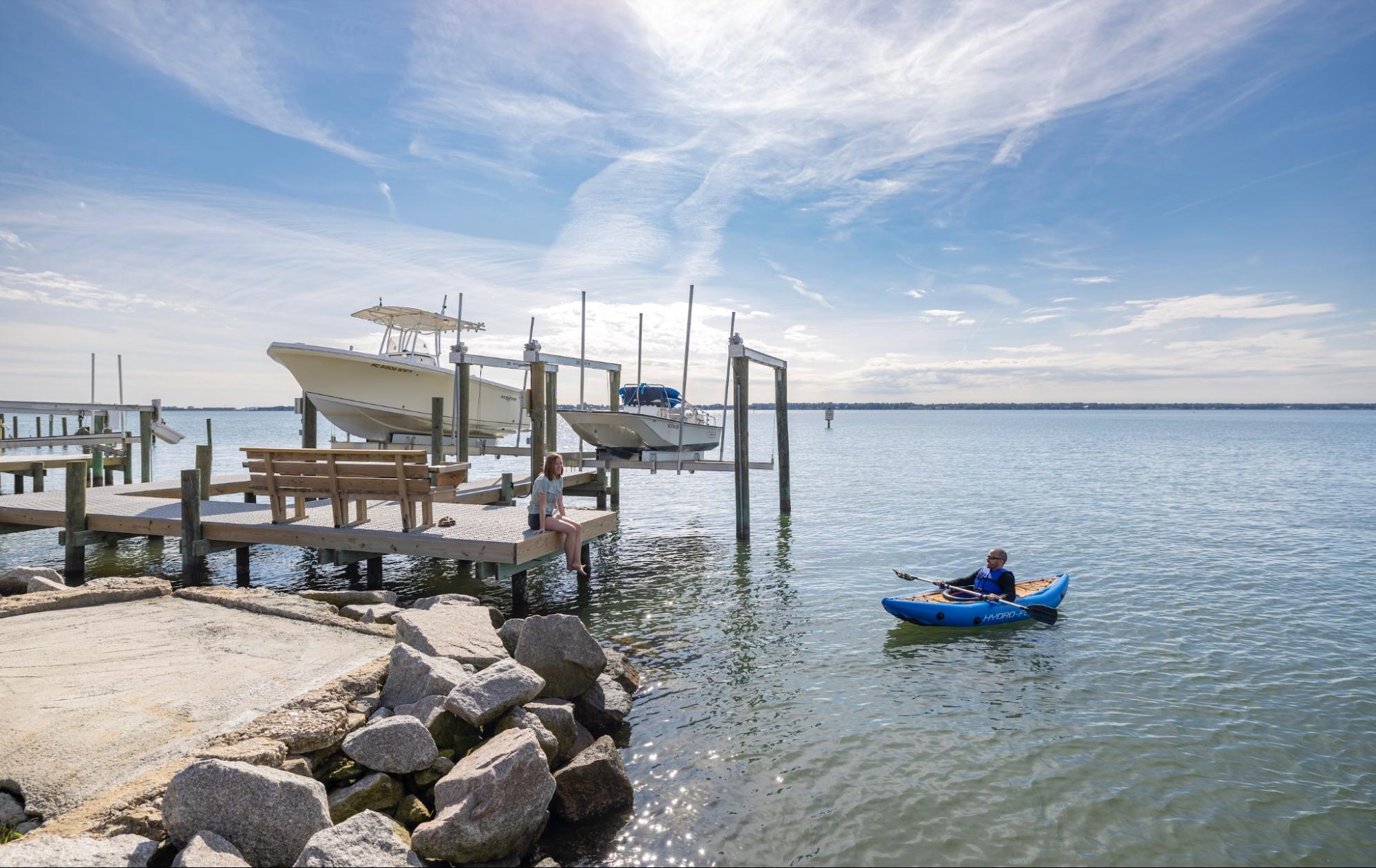 A person in a blue kayak is paddling toward a boat dock where another person is sitting. There are two boats on boat lifts next to the dock, and a wooden bench for sitting on the dock.