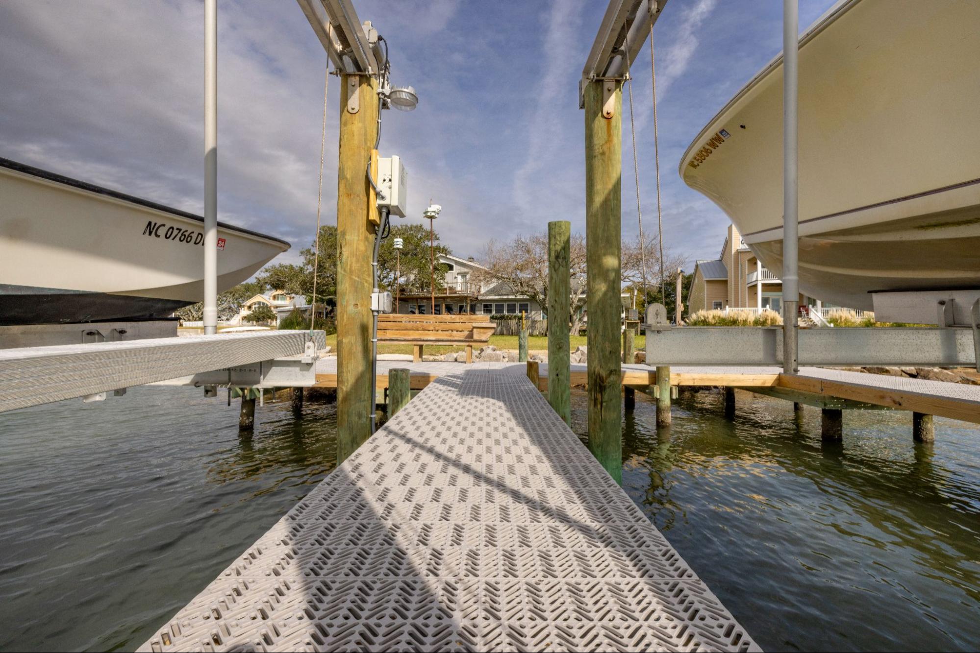 This image is from the perspective of standing on the end of a dock towards a bench and a house in the distance. There are two raised boats surrounding the dock, above the water.