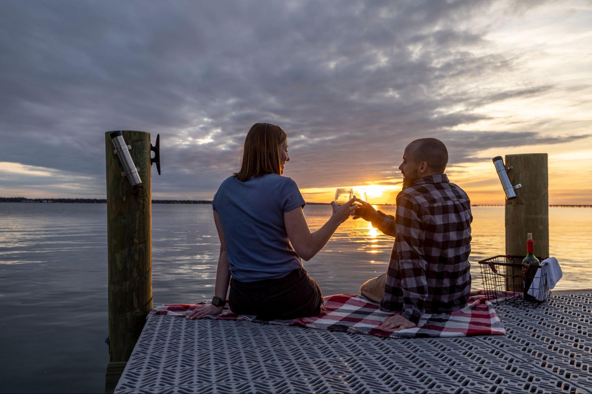 Two people are having a picnic on a dock at sunset. They are holding wine glasses and sitting on a blanket. 