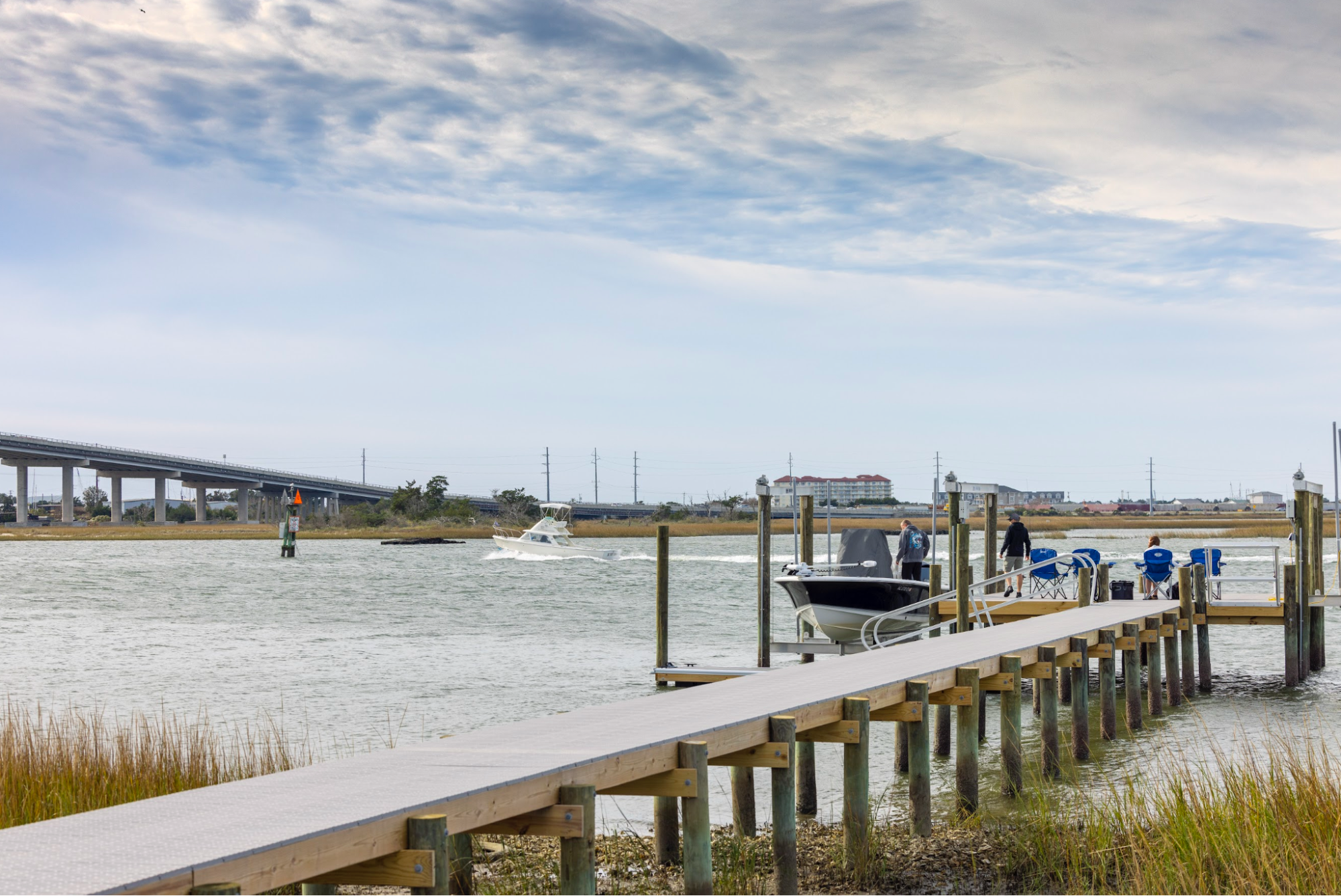 A family is getting into a boat from their dock and there is another boat passing by in the water. A commercial building is seen in the background, along with a highway that crosses over the water nearby. 