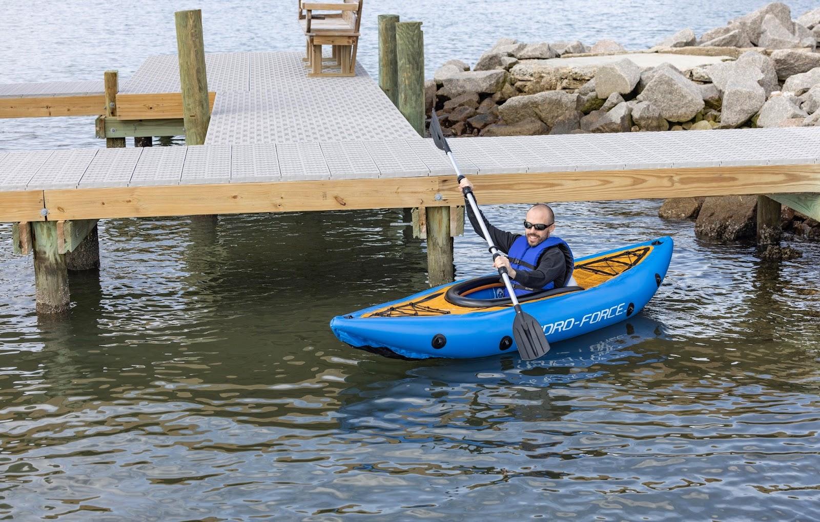 A person wearing sunglasses and a blue life vest is rowing in a blue kayak near a boat dock. 