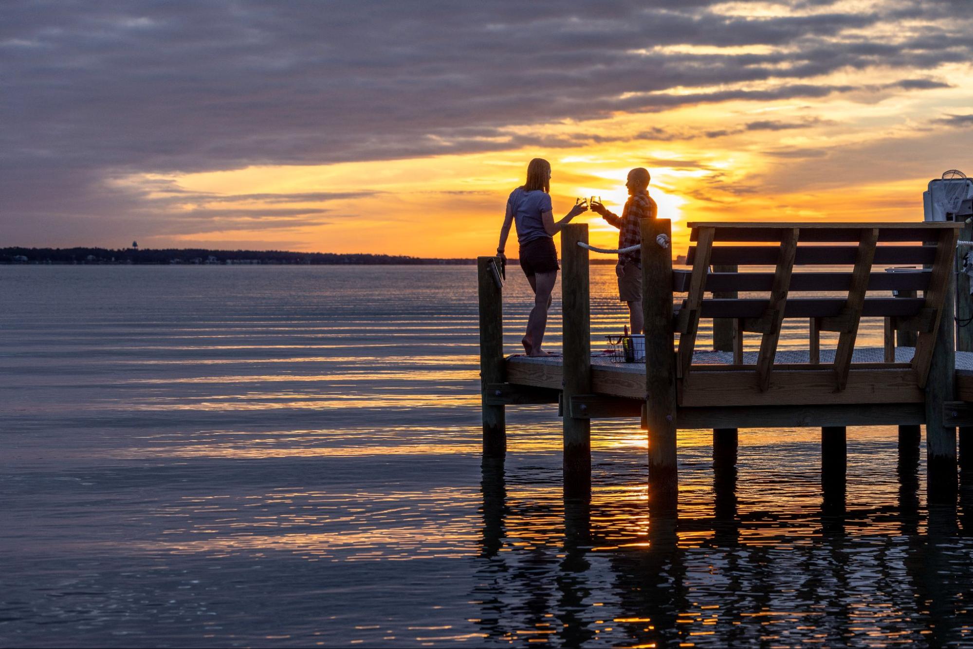 Two people cheersing at the end of a dock at sunset.