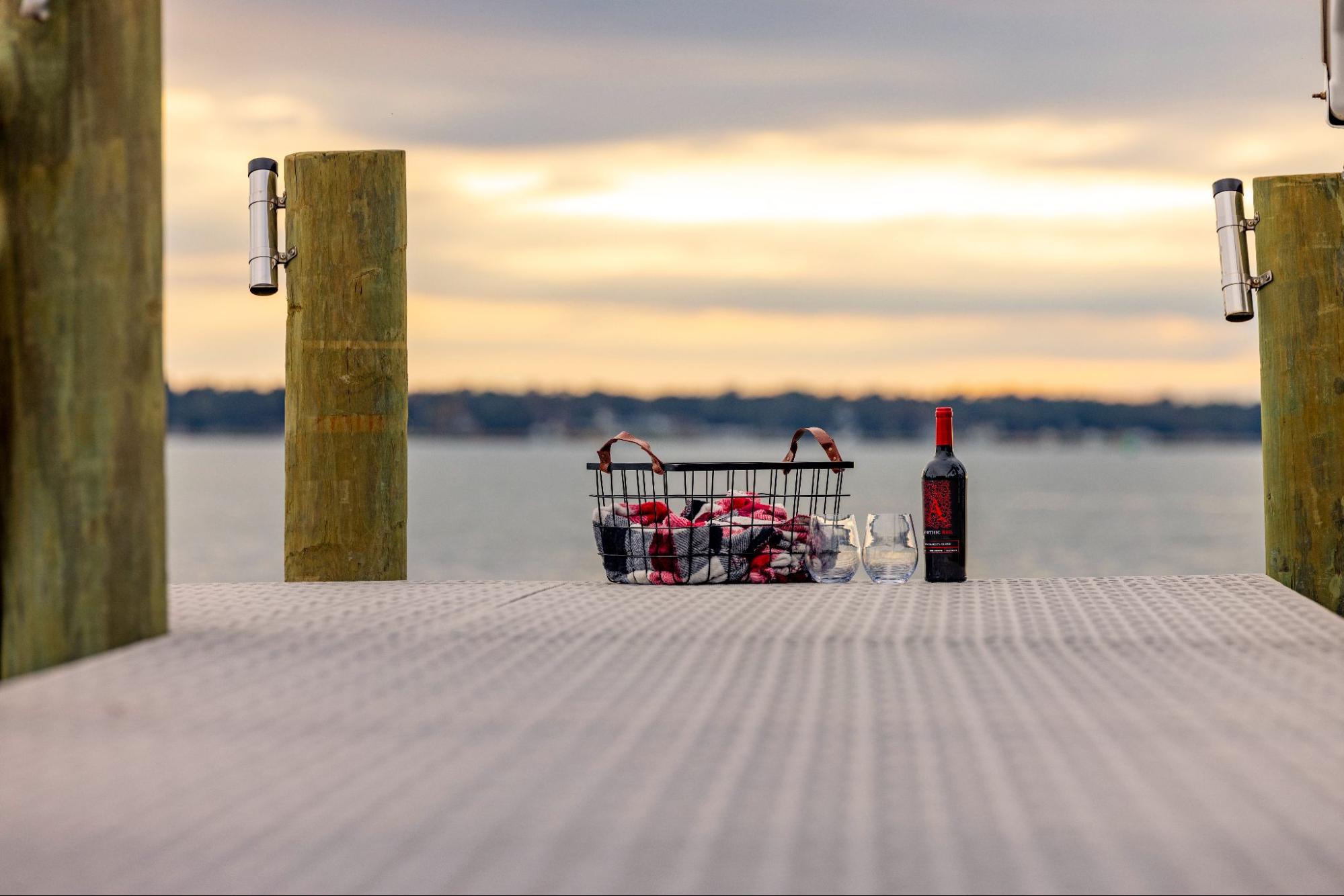 The photo depicts a low-angle close-up shot of a picnic basket with a bottle of wine and two glasses at the end of a dock. The dock is gray and there are three wooden pillars attached, two with fishing rod holders. 
