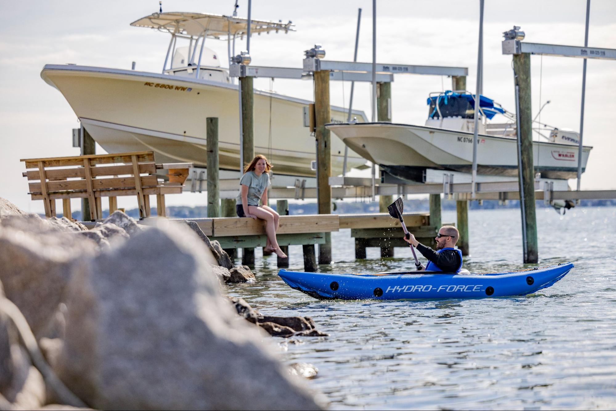 A person in a blue kayak approaches a dock, where there is another person sitting on the edge of the dock. There are two boats in the background suspended in boat lifts.