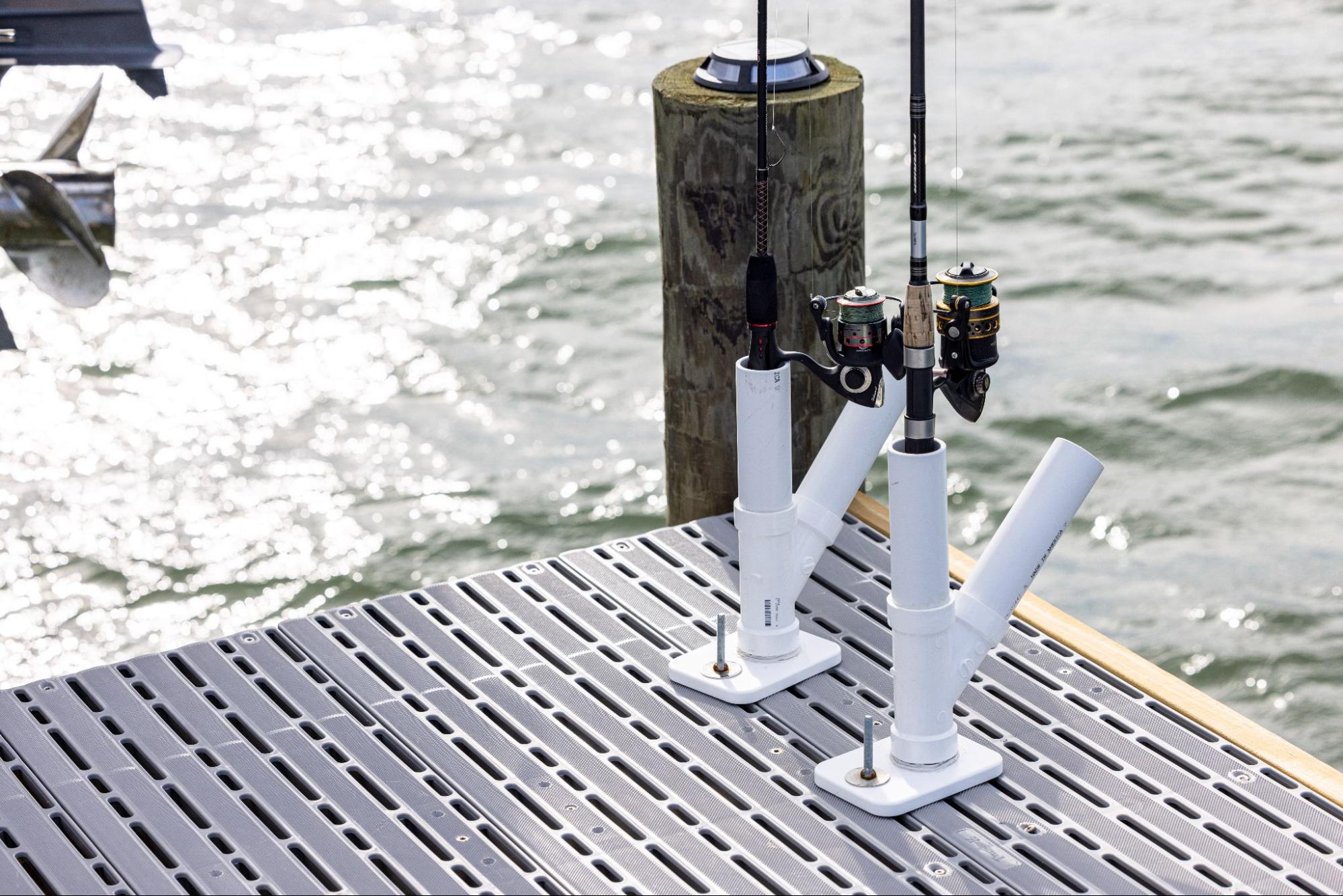 The photo depicts a close-up shot of two dock-mounted fishing rod holders. A wood pillar is attached at the corner of the dock and a body of water is in the background. 