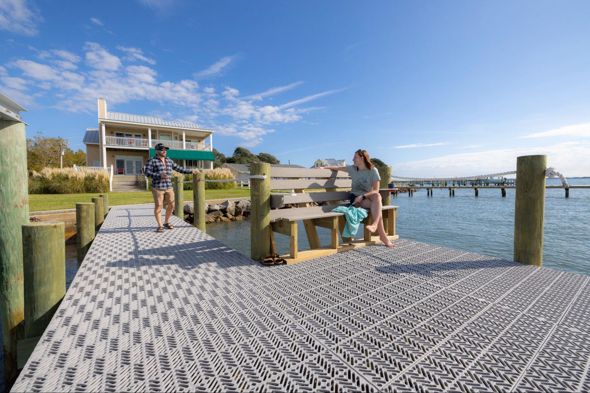 The photo is of a gray L-shaped dock with a bench and wood pillars attached at various points and a yellow house in the background. There are two people conversing on the dock and one person is seated on the bench and the other person is standing. 