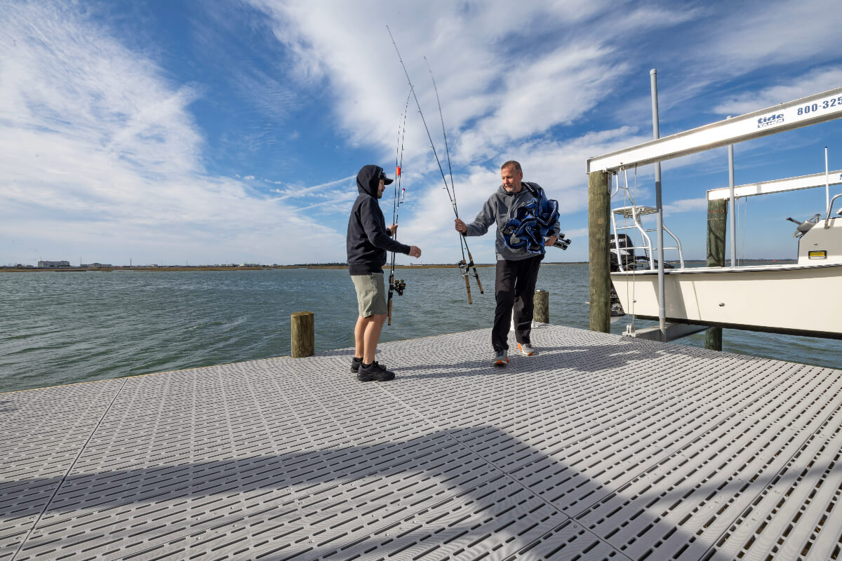 Two people are holding fishing rods while standing on a Titan Deck dock. There is a boat on a lift to the side of them. 