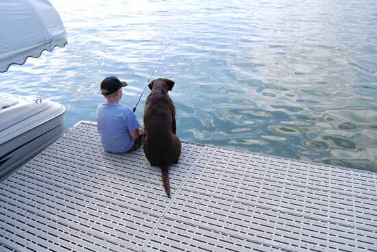 A young boy sits on a dock next to a large brown dog, both facing the water. The boy is wearing a baseball cap and holding a fishing pole. The water is calm in front of them and part of a boat is on the left side of the dock.